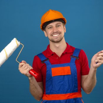 joven-constructor-uniforme-construccion-casco-seguridad-rodillo-pintura-pincel-gran-sonrisa-cara-sobre-pared-azul-aislada